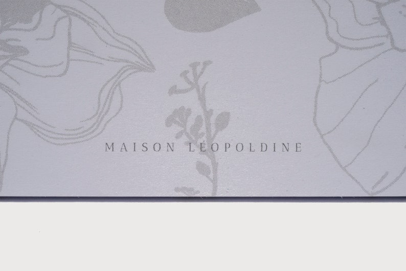 以下が含まれることがあります： 薄いグレーの背景に、花柄のデザインと「MAISON LEOPOLDINE」というテキストが銀色のフォントで書かれています。