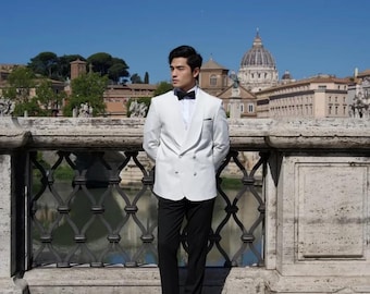 May include: A man wearing a white tuxedo jacket with a black bow tie, black pants, and black loafers. He is standing on a bridge with a view of a city in the background.