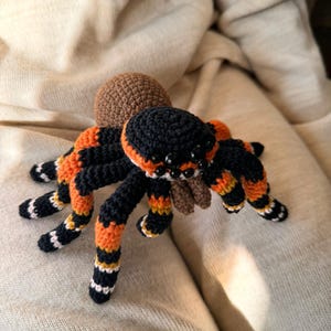 Puede incluir: Un juguete de araña de crochet con rayas negras, naranjas y blancas. La araña tiene una cabeza marrón y ocho patas.