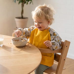 Könnte beinhalten: Ein Kind mit einer gelben Latzhose und einem gemusterten Langarmshirt sitzt an einem Holztisch und isst Müsli aus einer Schüssel. Ein Glas Milch und eine Tasse stehen ebenfalls auf dem Tisch, zusammen mit einer Obstschale.