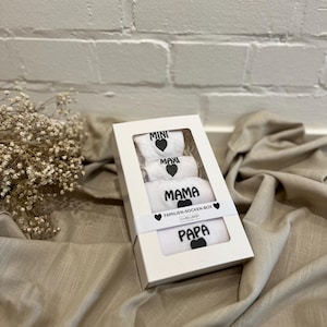 Puede incluir: Una caja de regalo blanca que contiene tres pares de calcetines blancos. Cada calcetín tiene un corazón negro y una palabra: "MINI", "MAXI", "MAMA" y "PAPA". La caja está sobre una superficie de tela beige.
