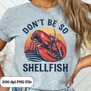 May include: A light gray t-shirt with the text "DON'T BE SO SHELLFISH". The design features a red lobster illustration set against a sunset background. The words are in a dark blue font.