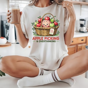 May include: Cream-colored t-shirt featuring a whimsical illustration of a brain in a basket of apples. The text "APPLE PICKING & OVERTHINKING" is below the graphic. The design includes red and green apples, and a tag that says "FRESH PICKED THOUGHTS".