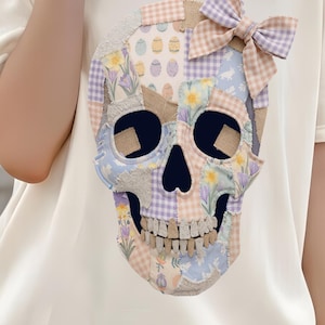 Könnte beinhalten: Cremefarbenes T-Shirt mit einem Patchwork-Schädel-Design und einer Schleife. Der Schädel besteht aus verschiedenen Pastellmustern, darunter Blumen- und Vichy-Drucke. Der Text "Visual Sensei" ist unten links aufgedruckt.