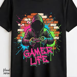 Puede incluir: Camiseta negra con una figura encapuchada sosteniendo un mando de videojuegos, sobre un fondo de pared de ladrillo con salpicaduras de pintura de neón. Las palabras "GAMER LIFE" se muestran en un estilo graffiti rosa y verde vibrante.