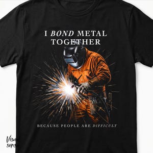 以下が含まれることがあります： 黒いTシャツ。オレンジ色のつなぎを着た溶接工が金属を溶接しているグラフィック。画像の上には「I BOND METAL TOGETHER」、下には「BECAUSE PEOPLE ARE DIFFICULT」の文字。
