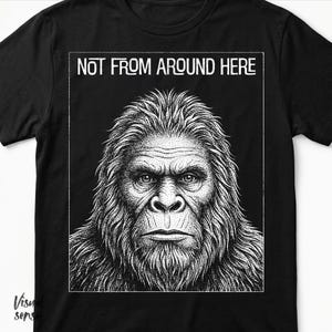 Pode incluir: Camiseta preta com uma ilustração detalhada em preto e branco do rosto de um Sasquatch. O texto "NOT FROM AROUND HERE" é impresso em branco acima da ilustração. O design é centrado na camiseta.