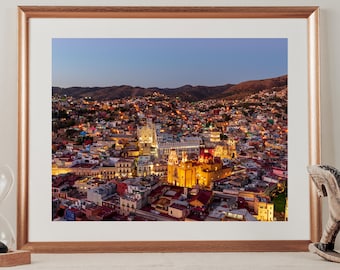 May include: An aerial view of a city at dusk, with a large church in the center. The city is lit up with lights, and the church is illuminated with warm, golden light. The sky is a soft blue, and the hills in the distance are silhouetted against the sky.