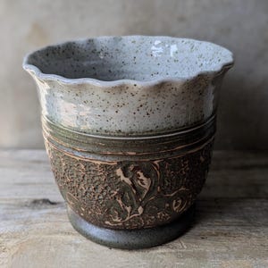 May include: A handmade ceramic plant pot with a speckled, light grey rim and a textured, dark brown lower section. The pot features a decorative floral design and sits on a small, dark grey base. The overall aesthetic is rustic and artisanal.