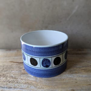 May include: A cylindrical ceramic bowl with a white interior and a blue exterior. The blue exterior features a band of alternating black and blue circles. The bowl sits on a wooden surface.