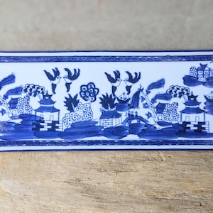 May include: Rectangular blue and white ceramic tray with a Willow pattern design. The tray features a detailed landscape scene with pagodas, trees, and birds. The border is a simple blue line. The tray is displayed on a wooden surface.