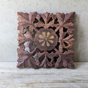 May include: A square, intricately carved wooden wall decor piece. The dark brown wood features a central floral design with a gold-coloured centre, surrounded by detailed leaf patterns. The piece is approximately 25 cm square.