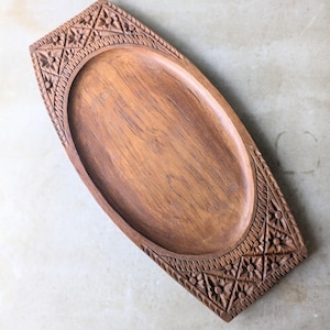 May include: A rectangular wooden tray with a smooth, oval-shaped recessed centre. The two shorter ends feature intricate hand-carved details, including a geometric grid pattern filled with small floral motifs and a textured border along the edges.