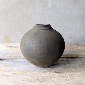 May include: A dark grey ceramic vase with a rounded body and a narrow neck. The vase has a textured surface and horizontal lines near the top. It is displayed on a weathered wooden surface.
