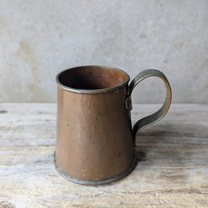 May include: A small, hammered copper mug with a handle. The mug has a slightly rounded bottom and a flared top.