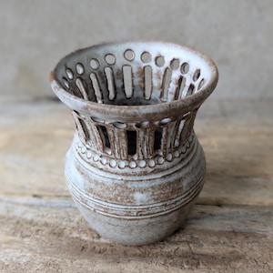 May include: A small, off-white ceramic vase with a textured surface and a perforated design. The vase has a wide, open top with evenly spaced holes and vertical slots around the body. The vase is approximately 13 cm tall and has a rustic, handmade appearance.