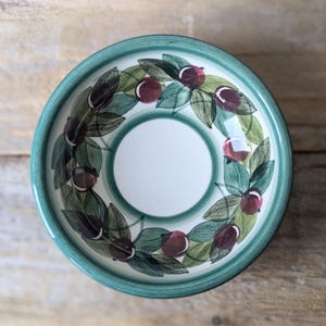 May include: A round ceramic bowl with a teal rim and a white interior. The bowl is decorated with a wreath of green leaves and maroon berries. The bowl is set against a wooden background.