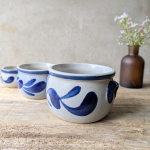 May include: Three round ceramic pots with a light grey speckled finish and a dark blue hand-painted leaf pattern. Each pot features a thick blue rim and a small handle on the side. The pots are arranged in a line, decreasing in size from front to back.