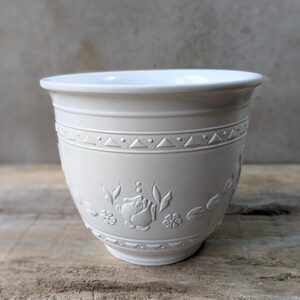 May include: Cream-coloured ceramic plant pot with a rounded body. The pot has embossed floral and geometric patterns around the sides. The rim is smooth and the interior is white. It sits on a wooden surface.