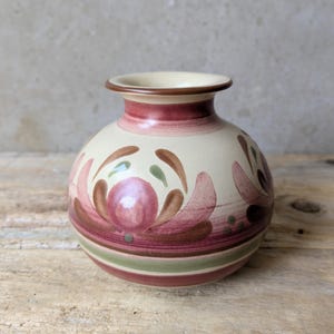 May include: A round ceramic vase with a cream-coloured base featuring hand-painted floral motifs in shades of dusty rose, sage green, and brown. The vase has a short, flared neck with a brown rim and horizontal bands of colour encircling the lower body.