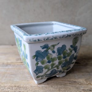 May include: A square, white ceramic bonsai pot with a blue and green floral design. The pot has a slightly flared rim and small feet. The floral pattern includes leaves and vines. The pot is sitting on a wooden surface.