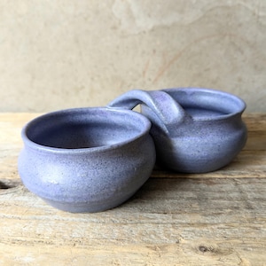 May include: A pair of connected ceramic bowls with a speckled lavender glaze. The bowls feature rounded bodies, wide openings, and a single arched handle bridging the two vessels, creating a unified piece with a smooth, consistent finish.