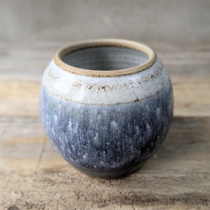 May include: A handmade ceramic bowl with a rounded form. The bowl displays a colour gradient, shifting from white at the top to shades of blue and grey towards the base. The rim has a natural, unglazed finish.
