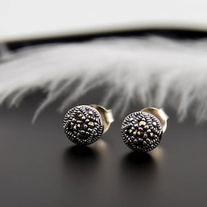 Könnte beinhalten: Ein Paar silberne Ohrstecker. Jeder Ohrring hat ein rundes, strukturiertes Design mit dunkler, oxidierter Oberfläche und kleinen, funkelnden Akzenten. Die Ohrstecker sind auf einer schwarzen Oberfläche mit einer weißen Feder im Hintergrund platziert.