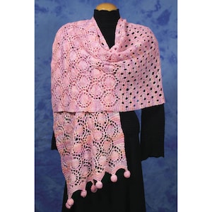 May include: A pink crocheted shawl with a lacy pattern and pom-poms on the fringe. The shawl is draped over a mannequin wearing a black mock neck shirt.