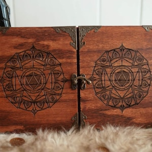 Puede incluir: Una caja de madera con un pestillo y diseños grabados ornamentados en la parte delantera y trasera. Los diseños presentan un mandala con un dado de 20 caras en el centro.