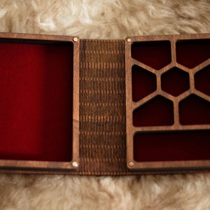 Op de afbeelding: Open houten doos met een rode fluwelen binnenkant. De doos is verdeeld in compartimenten met een geometrisch ontwerp aan &eacute;&eacute;n kant. De doos is open, waardoor de binnenkant zichtbaar is, en staat tegen een zachte, lichtgekleurde achtergrond.