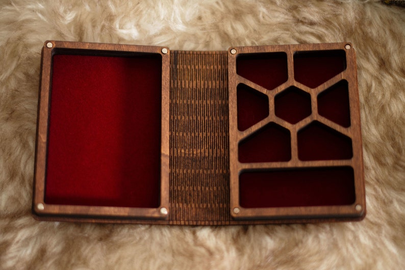 Op de afbeelding: Open houten doos met een rode fluwelen binnenkant. De doos is verdeeld in compartimenten met een geometrisch ontwerp aan &eacute;&eacute;n kant. De doos is open, waardoor de binnenkant zichtbaar is, en staat tegen een zachte, lichtgekleurde achtergrond.