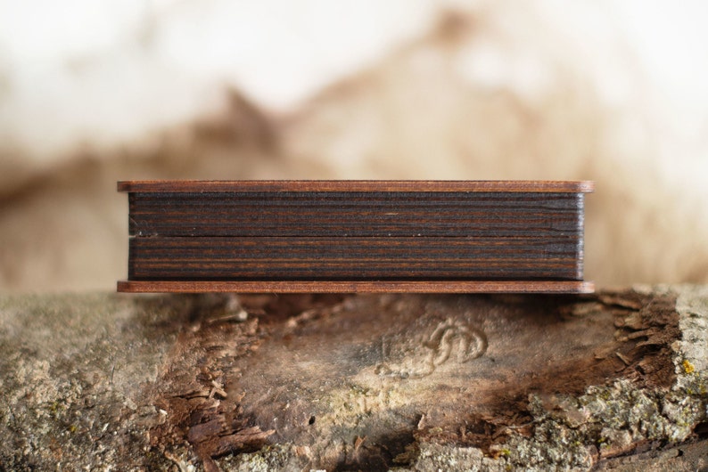 Op de afbeelding: Een kleine, rechthoekige houten doos met een donkerbruin interieur en een lichtere bruine lijst. De doos rust op een getextureerd, natuurlijk houten oppervlak, met een vage achtergrond.