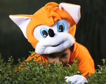 May include: A child wearing an orange and white fox costume is hiding behind a green bush. The costume has a large, white head with blue eyes and a smiling mouth.