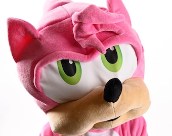 May include: Pink plush mascot costume with a white face, green eyes, and a black nose. The costume has a pink hood with a white lining and a white shirt underneath.