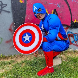 May include: A person in a Captain America costume, featuring a blue jumpsuit, mask, and shield. The shield is red, white, and blue with a star. The costume includes red boots and gloves. The background is a graffiti-covered wall and green grass.