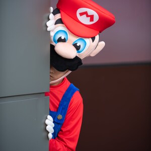 Op de afbeelding: Een persoon verkleed als Mario uit de Super Mario-videogameserie kijkt om een hoek. Het kostuum is rood en blauw met een witte bliksem op de hoed.