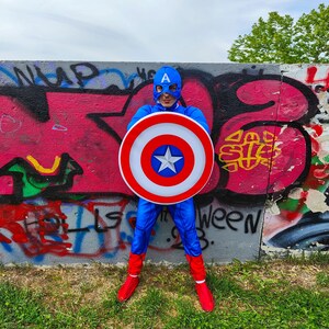 May include: A person in a Captain America costume, holding a shield with a white star on a blue background, surrounded by red and white concentric circles. The costume is blue with red boots and a blue mask with the letter "A". The background is a graffiti-covered wall.