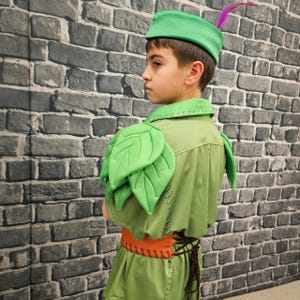 May include: A green costume with leaf-shaped shoulder pads, an orange belt, and a matching hat with a purple feather. The costume is designed with a medieval or fantasy theme, possibly for a play or cosplay.