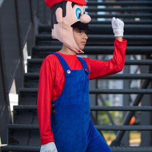 Op de afbeelding: Een kind verkleed als Super Mario, met een rode hoed met een witte M, een rood shirt met lange mouwen en een blauwe overall. Het kind staat op een metalen trap.