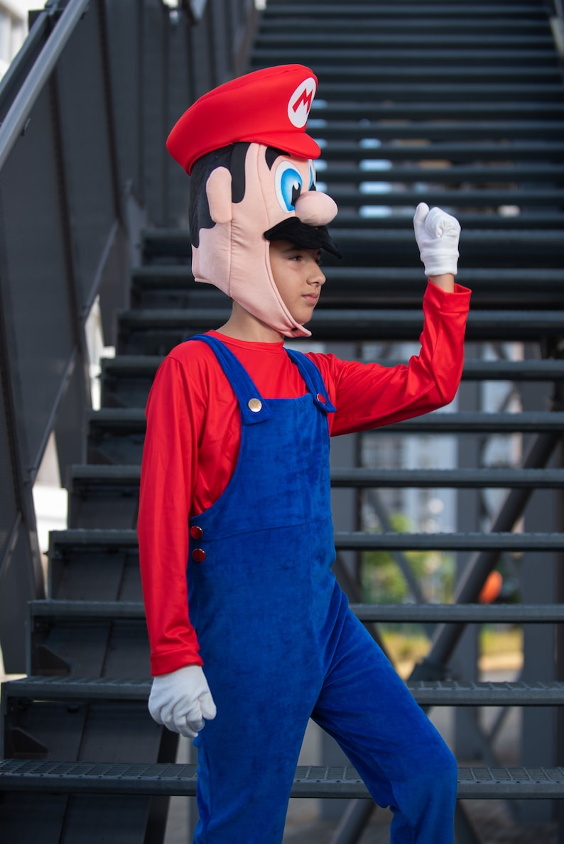 Op de afbeelding: Een kind verkleed als Super Mario, met een rode hoed met een witte M, een rood shirt met lange mouwen en een blauwe overall. Het kind staat op een metalen trap.