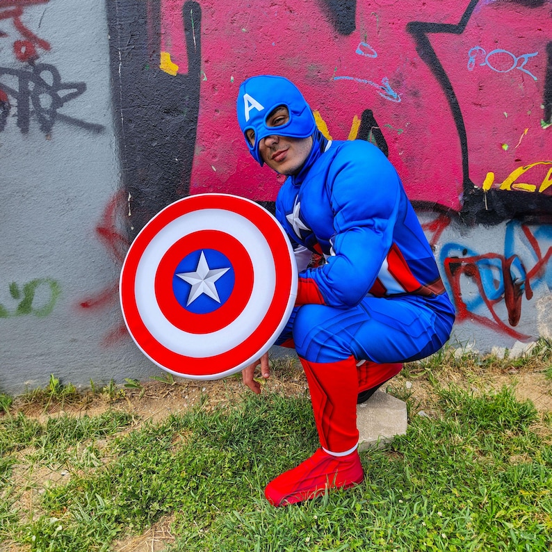 May include: A person in a Captain America costume, featuring a blue jumpsuit, mask, and shield. The shield is red, white, and blue with a star. The costume includes red boots and gloves. The background is a graffiti-covered wall and green grass.