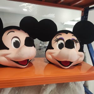 Pode incluir: Duas máscaras de cabeça de personagem de pelúcia com grandes orelhas pretas redondas, rostos cor de pêssego, narizes pretos e sorrisos largos. Uma máscara tem olhos simples, enquanto a outra apresenta cílios pretos longos e sombra roxa.