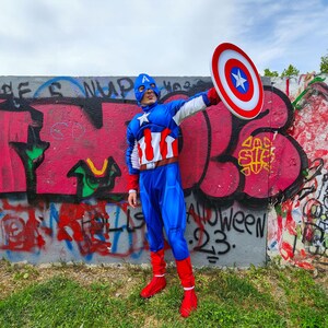 May include: A person in a Captain America costume, holding a shield. The costume is blue, red, and white, with a star on the chest and a blue mask. The shield is red, white, and blue. The background is a graffiti-covered wall.
