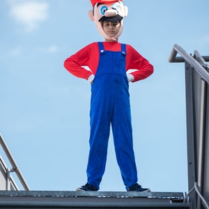 Op de afbeelding: Een kind verkleed als Super Mario, met een rode hoed met een witte 'M' en een rood shirt met een blauwe overall. Het kind staat op een metalen trap tegen een blauwe lucht.