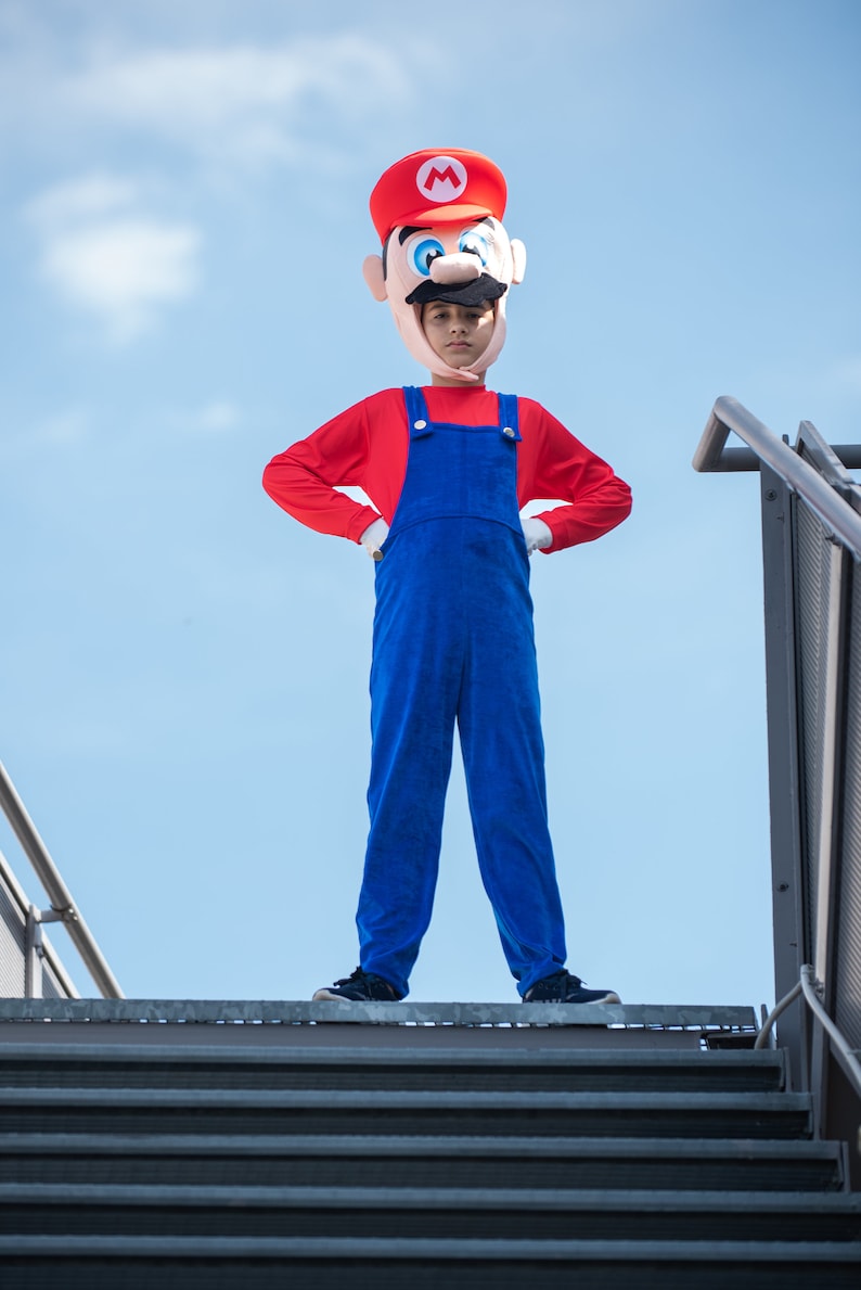 Op de afbeelding: Een kind verkleed als Super Mario, met een rode hoed met een witte 'M' en een rood shirt met een blauwe overall. Het kind staat op een metalen trap tegen een blauwe lucht.