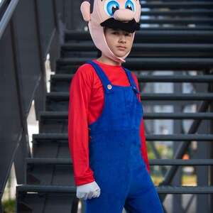 Op de afbeelding: Een kind verkleed als Mario uit de Super Mario-videogameserie. Het kind draagt een rood shirt met lange mouwen, een blauwe overall en een rood-witte Mario-hoed. Het kind staat op een metalen trap.