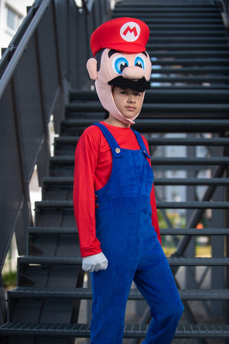 Op de afbeelding: Een kind verkleed als Mario uit de Super Mario-videogameserie. Het kind draagt een rood shirt met lange mouwen, een blauwe overall en een rood-witte Mario-hoed. Het kind staat op een metalen trap.