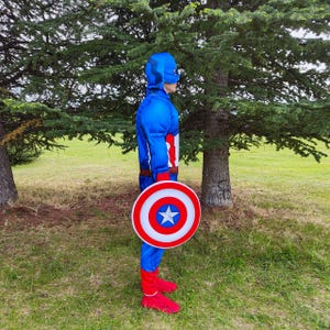 May include: A blue Captain America costume with a matching helmet and shield. The shield is red, white, and blue with a star in the centre. The costume includes red gloves and boots. The person is standing in a grassy area with trees.