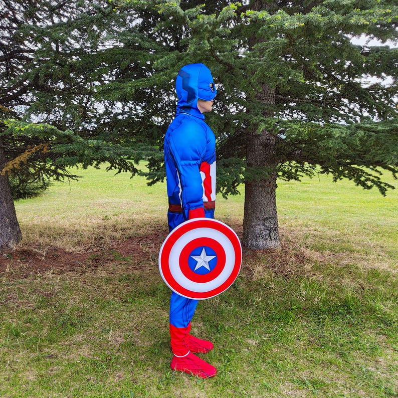 May include: A blue Captain America costume with a matching helmet and shield. The shield is red, white, and blue with a star in the centre. The costume includes red gloves and boots. The person is standing in a grassy area with trees.